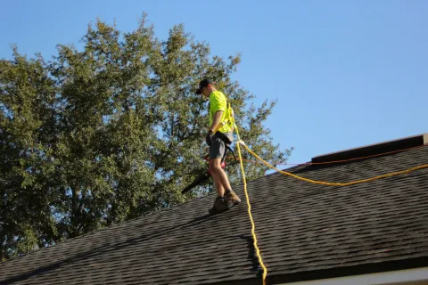 Worker in safety gear using blower to clean roof shingles on a sunny day with blue sky and trees nearby