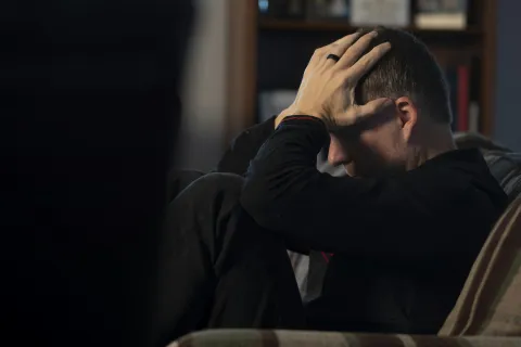 Man sitting on couch with hands on head appearing stressed or anxious in dimly lit room.