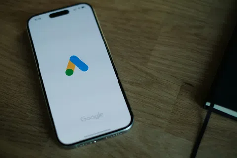 Smartphone displaying the Google Ads logo on a wooden table next to a closed notebook.