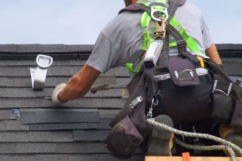 Roofer installing shingles on a house roof, showcasing safety gear and tools in action.
