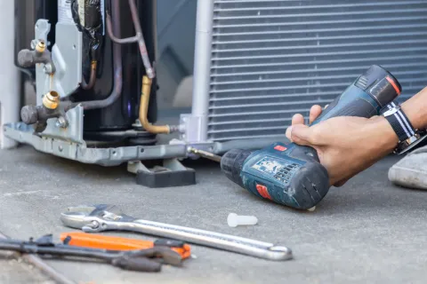 Technician fixing an air conditioning unit using a cordless drill and various hand tools on concrete floor.