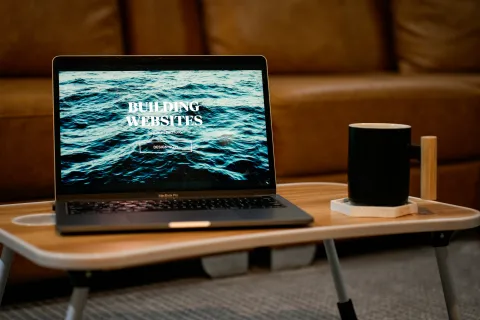 Laptop on wooden table displaying website with ocean waves and text building websites, next to black coffee mug.