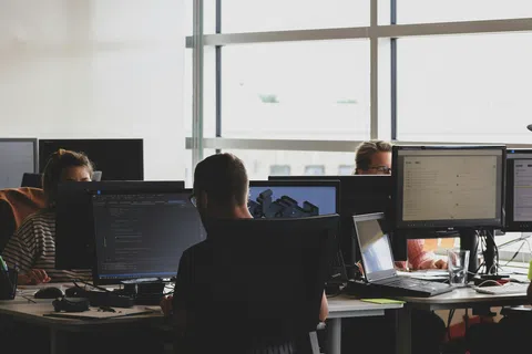 People working at desks with multiple computer monitors in a bright open office space.