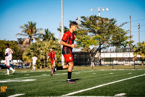 Soccer player in red and black uniform running on field during daytime match under clear blue sky.