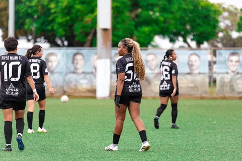 Women soccer players in black River Miami FC uniforms on a green field during a game or practice session.