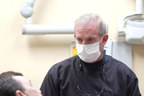 Dentist wearing a mask and black scrubs examining a patient in a dental clinic with equipment in the background