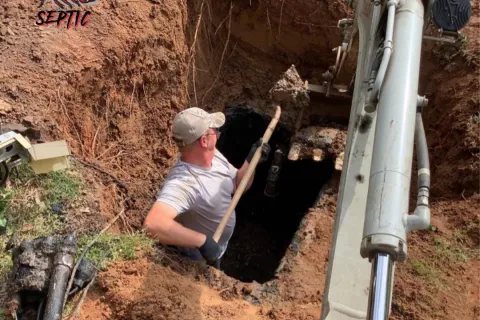 Worker shoveling dirt from a septic tank excavation site next to heavy machinery on a sunny day.