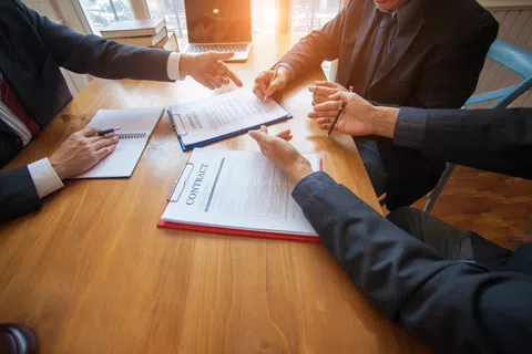 Business professionals discussing and signing contracts at a wooden table in a meeting room with sunlight.