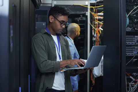 Technician using a laptop to manage servers inside a data center server room with network cables and equipment.