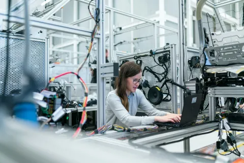 Woman working on laptop surrounded by automotive electronic components and wiring in a modern lab.