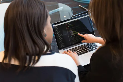 Two women collaborate on coding by reviewing and pointing at a laptop screen showing programming code.