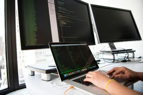 Hands typing on a laptop with programming code on screen and two large monitors displaying code in a bright office.