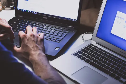 Hands typing on a Dell laptop keyboard next to an open MacBook Air on a desk with a light background