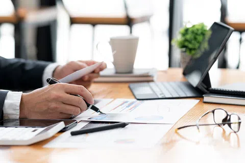 Person analyzing business charts with pen at a desk with laptop, tablet, glasses, and coffee cup.
