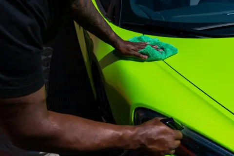 Person cleaning a vibrant green sports car with a microfiber cloth under bright lighting.
