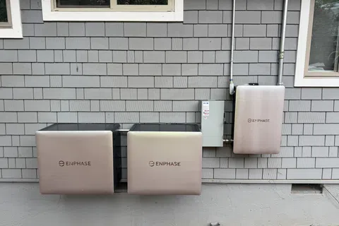 Three Enphase energy storage units mounted on a gray shingled exterior house wall under two windows.