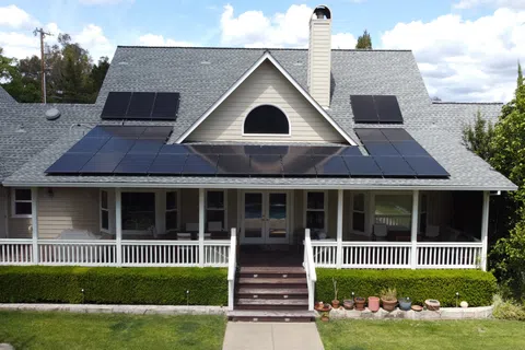 Suburban house with a gray roof featuring solar panels on a sunny day with a green lawn and blue sky.