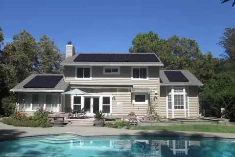 Two-story house with solar panels on the roof and a swimming pool in the backyard under a clear blue sky.
