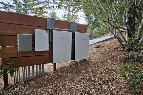 Tesla Powerwall home battery units installed on a wooden panel in an outdoor wooded area.