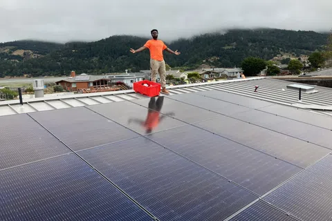 Person in orange shirt standing on a roof with solar panels under a cloudy sky.
