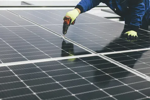 Technician installing solar panels using a power drill wearing gloves and blue workwear on a sunny day