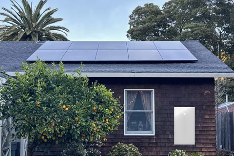 Solar panels on a house roof with a Tesla Powerwall battery mounted on the exterior wall beside a window and orange tree.