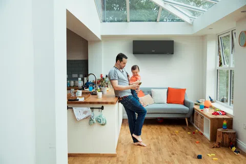Father and child using smartphone in bright living room with sofa, air conditioner, and toys on wooden floor.