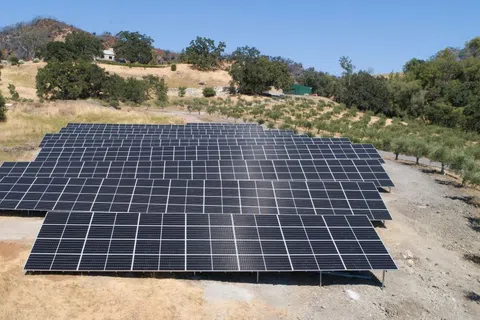 Aerial view of solar panels installed in a dry, grassy field surrounded by trees and hills.