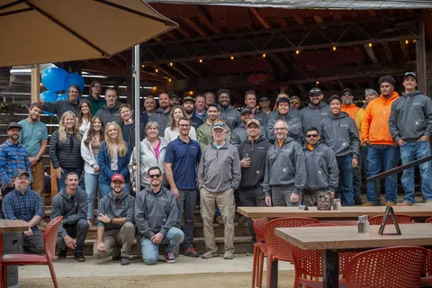 Group photo of a diverse team outdoors, smiling together under an awning at a lively event.