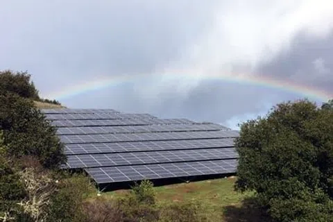 Solar panels on a green hillside with a rainbow and cloudy sky in the background.