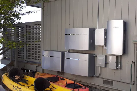 Home energy storage system with sleek silver batteries mounted on exterior wall above kayaks under tree shade.