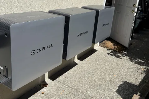 Three Enphase solar energy storage units mounted on a wall in outdoor sunlight near an open door.