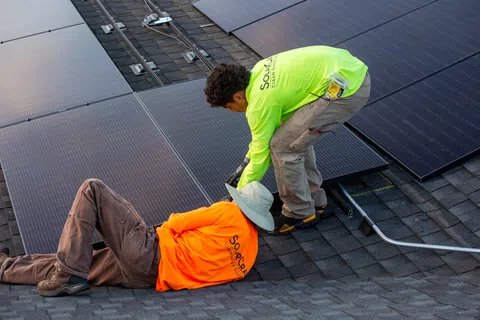 Two workers installing solar panels on a roof with safety gear and bright work shirts in daylight.