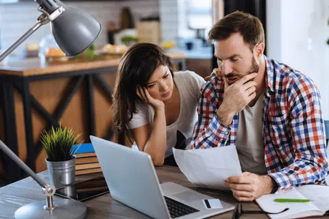 Couple reviewing bills and finances with laptop and documents at home, appearing concerned and focused