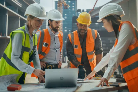 Construction team wearing safety gear reviewing plans and laptop at building site under development.
