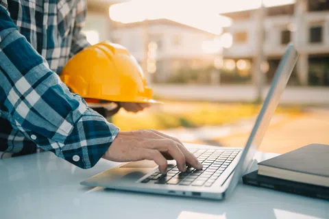 Engineer in plaid shirt holding a yellow safety helmet typing on a laptop at a construction site desk.