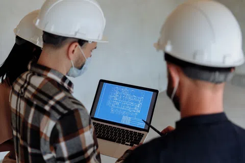 Engineers wearing white helmets reviewing architectural blueprint on a laptop at a construction site indoors.