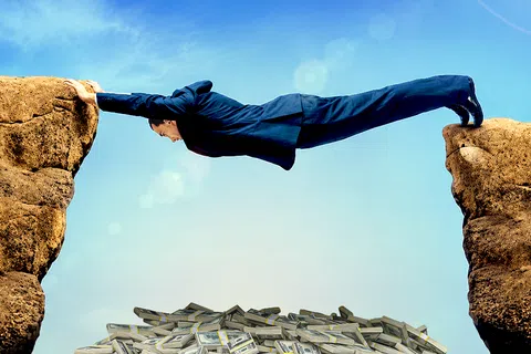 Businessman stretches across a rock gap above a pile of money under a clear blue sky.