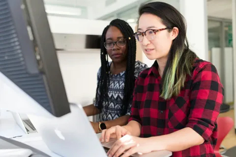 Two women collaborating on a laptop and desktop computer in a bright modern office setting.