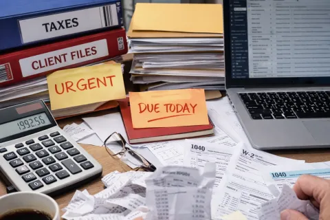 Desk cluttered with tax documents, calculator, laptop, urgent and due today notes, and client file folders.
