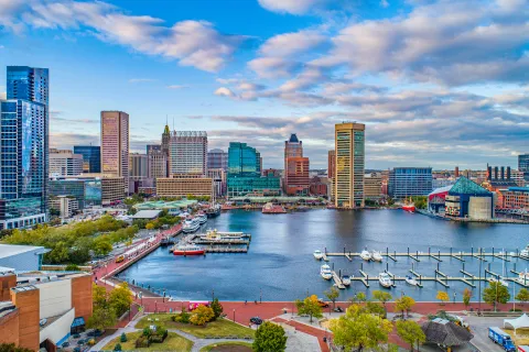 Baltimore Inner Harbor waterfront with boats, marina, city skyline, and cloudy blue sky in vibrant daylight.