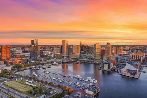Baltimore skyline at sunset with Inner Harbor, boats docked, and vibrant orange and purple sky above cityscape.