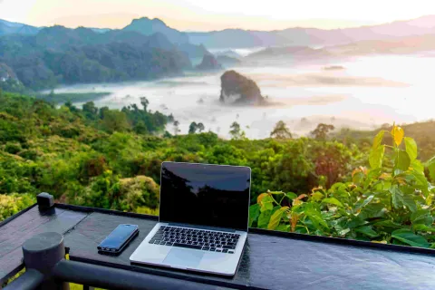 Laptop and smartphone on wooden desk overlooking misty green mountains at sunrise.