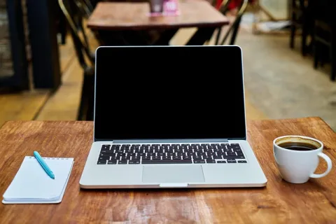 Laptop with blank screen, notebook with pen, and cup of coffee on wooden table in a cafe setting.