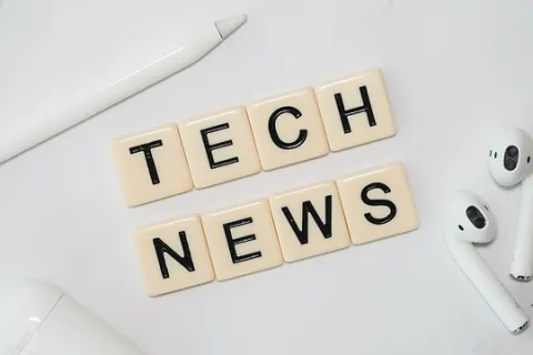 Tech news spelled with letter tiles next to white wireless earbuds, a pen, and charging case on a white surface.