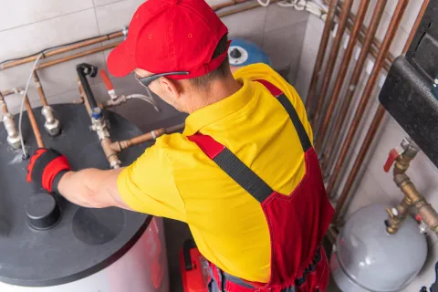 Repairman in overalls, hat, and gloves repairs water heater