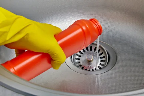 pink gloved hand pours red chemical cleaner down drain