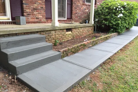 Newly poured smooth gray concrete steps and walkway alongside a red brick house with garden area.