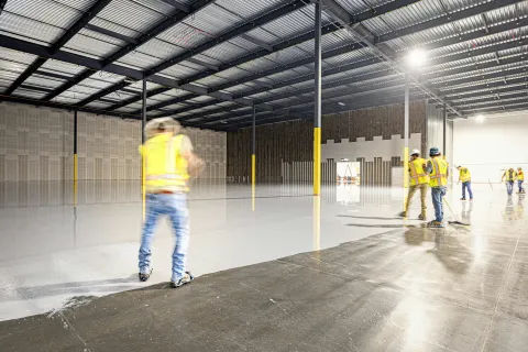 Construction workers applying a smooth coating to a large warehouse floor under a steel roof.