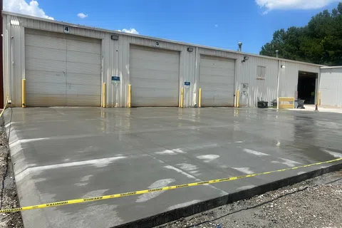 Freshly poured concrete slab in front of industrial garage doors with caution tape around the site under blue sky.